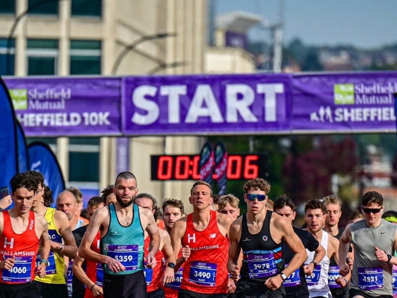 Runners in a Sheffield 10K event passes barriers lined with colourful sponsor banners. The runners head towards the camera from the start line.
