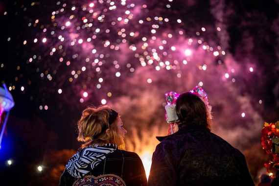 Two people wearing fancy head gear stand watching the dark sky lit up with purple fireworks