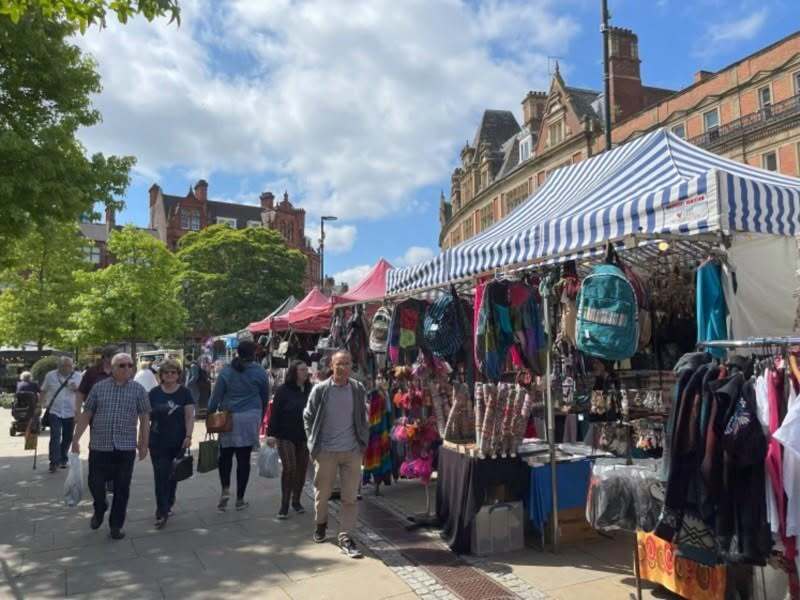 People walk past colourful market stalls in Sheffield. There are trees and tall buildings in the background, with a cloudy sky overhead.