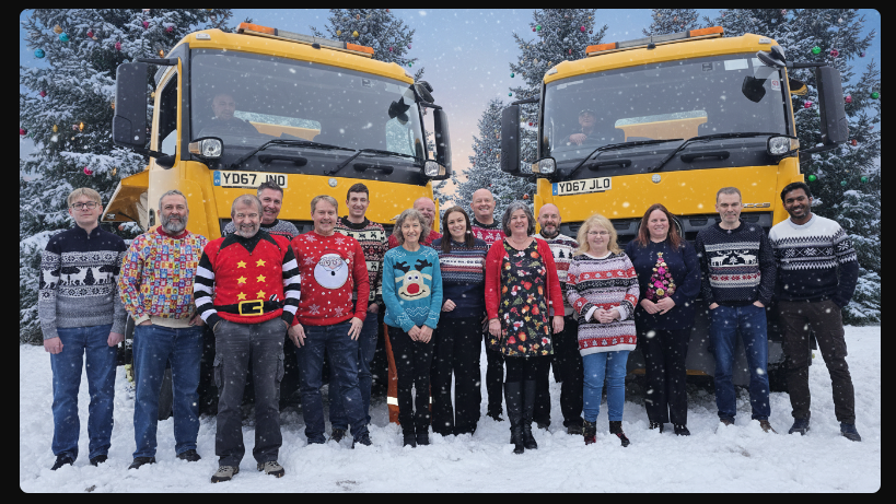 Roads staff in wearing festive jumpers in front of gritters and an edited snowy background