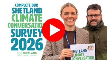 Climate Officers Elliot and Anne hold a copy of the Shetland Climate Conversation survey and smile at the camera 