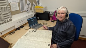 A woman sits in front of a desk with a historic newspaper open in front of her. 