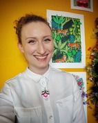 a woman with a wide smile and red curly hair tied back. She is wearing a white shirt with a necklace with a pink flower pendant