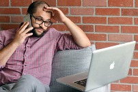 man holding his head staring at his phone and laptop