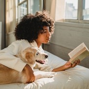 Woman reading a book, her dog is next to her and her arm is around the dog.