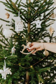 Close up of a real Christmas tree with lights and decorations on it and a hand hanging a decoration up.