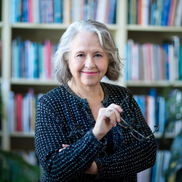 Canadian author Lesley Crewe. She is looking at the camera and has her eyeglasses in her hand, behind her there are bookshelves filled with book.
