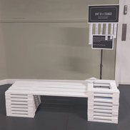 A small white bench made from wood. A sign behind it reads 'Don't be a stranger'.
