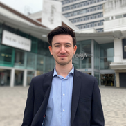 A young man in his 20's with dark hair smiling outside of the Victoria Centre. He is wearing a navy suit jacket over a light blue shirt.