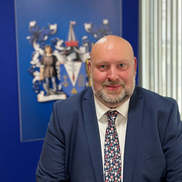 Cllr Tony Cox wearing a navy blue suit jacket, white shirt and a pink, blue and white flowery tie. He is sitting in front of the Council Coat of Arms.