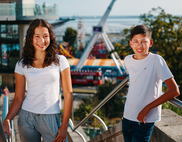 Two teenagers smiling at the top of the steps that lead down to the seafront. Some of the rides can be seen in the background out of focus.