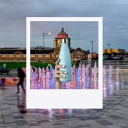 Hand with fingernails painted white holding a pale blue reusable water bottle in front of the seafront fountains and the Kursaal in the background.