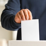 A close up of a man wearing a blue jumper. His hand is holding an envelope and putting it into a ballot box.