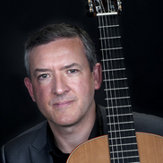 A head shot of guitarist Gary Ryan. He is wearing a dark suit jacket with the neck of a guitar resting on his shoulder.