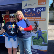 Two women dressed in navy blue T-shirts that say Could you Foster? They are standing in front of a supporting roller banner.