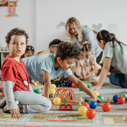 Children playing on a floor mat. There are toys scattered around and two adults in the background.