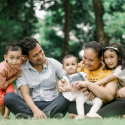 A family of five of Asian heritage. They are sitting outdoors on the grass and smiling at each other.