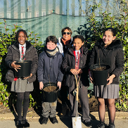 4 school children stand in front of a ward councillor. 3 of are holding tree saplings in a planter.