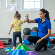A small child, hand-in-hand with a blue-clad physiotherapist, navigates a path of vibrant, plastic stepping stones.