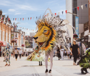 A vibrant parade surges down Southend High Street, where a participant is wearing an enormous lifelike lion's head.