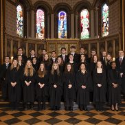 The choir of King's College, all dressed in black academic robes stood inside an ornate chapel. There are stained glass windows behind them.