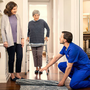A nurse moves a rug at the home of an elderly person using a walking aid, who stand smiling in the doorway of a hallway.