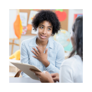 A Black-British woman with curly dark hair and green eyes talking to another person. She is holding a clipboard and has one hand on her chest.