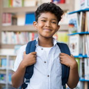 A boy wearing a school uniform and holding his backpack from his shoulders is smiling at the camera.