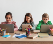 Three children seated at the same table with open laptops in front of them. There are pens and pencils on the wooden table.