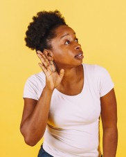 A Black British lady against a yellow wall leaning to one side with her hand over her ear as if listening carefully