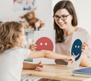 A woman in front of a child showing poster with a blue smiley face in one hand and a red sad face on the other.
