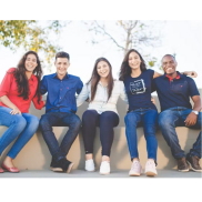 A group of 5 people outdoors. They are sitting on a wall with their arms on each other and smiling at the camera.