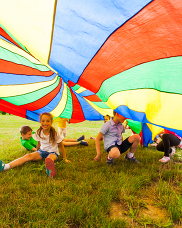 Children playing outside under a rainbow parachute.