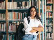 A female student with a backpack and arms crossed against a bookshelf.