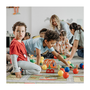 A group of children playing on a floor mat. There are toys scattered around and two adults in the background.