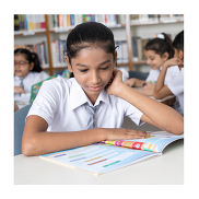 A girl in a school uniform sitting at a table reading a book. She is in the school library. There are bookshelves and two pupils in the background.
