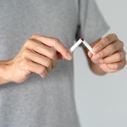 A close up of the mid section of man wearing a grey t-shirt breaking a cigarette in half with his hands.