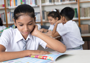A school girl reading a book in the school's library. There are bookshelves and other pupils in the background.