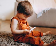 Toddler on an orange dungaree sitting on a light brown carpet looking at a white object in his hands.