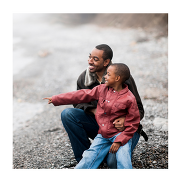A smiling man and a young boy of Black British ethnicity sit together outdoors on a pebble beach.