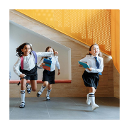 School children running in a school hall. They are wearing uniforms and carrying their backpacks.