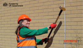 A women wearing ppe swinging a sledgehammer at a brick wall