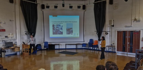 Two people stand in a school hall, showing a large screen displaying food hygiene information