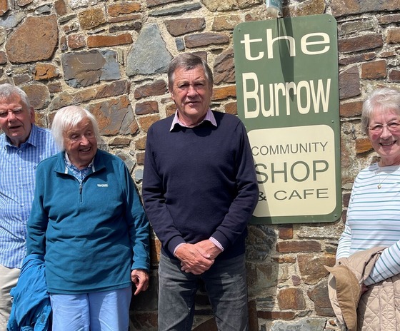 Four people stand next to a traditional stone wall and a green sign with cream writing of 'The Burrow Community Shop and Cafe