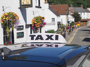 Taxi sign on a vehicle parked in a village street lined with hanging flower baskets, shops and residential buildings.