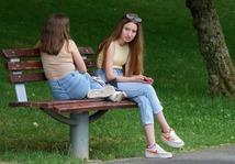Two teenage girls on a bench wearing jeans and vest tops, left one with her back to the camera and legs on bench, right one with mobile phone in hand