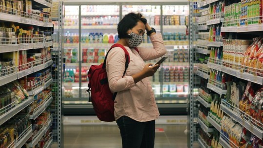 A woman stands in a supermarket aisle with her phone in her hand. She appears stressed.