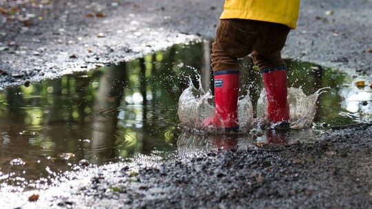 A small child, seen from behind, wearing a yellow raincoat and red wellies, jumping in a puddle