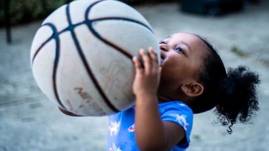 A little girl of about 3 years old holds a netball/basketball which is nearly bigger than her