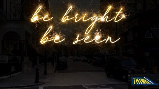 The words 'be bright be seen' handwritten in bright, glowing letters across an image of a murky street
